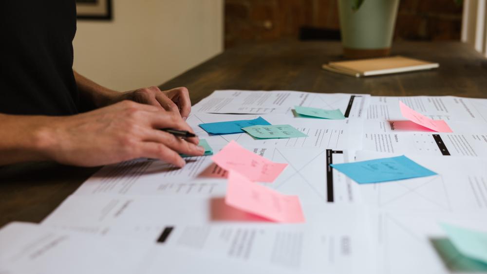 desk with diagrams and post it notes on. A person is stood next to the desk, only their hands are visible