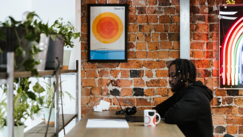 Person at a desk on a laptop, with plants on the left and a neon sign on the right 