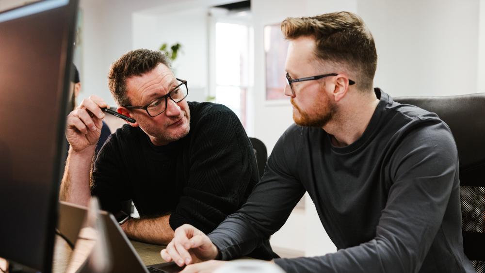 Two people talking, one typing on a laptop