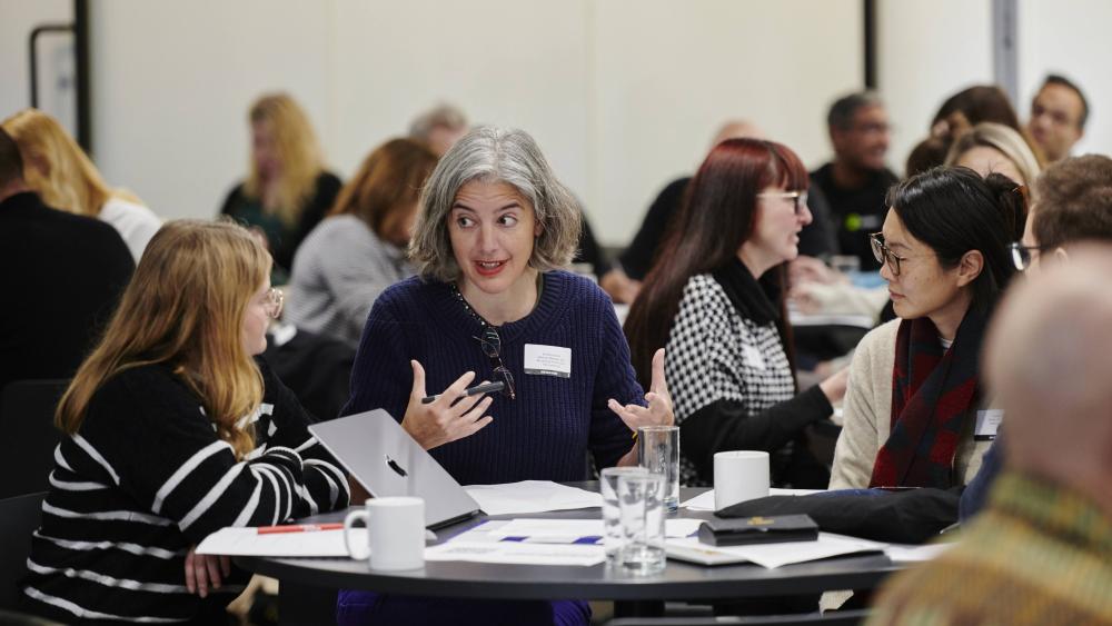 a busy conference, people are sat around a table in the foreground, with many more people sat on tables in the background
