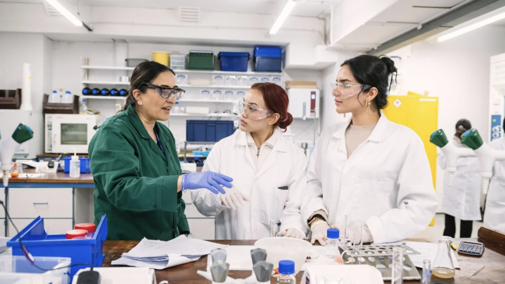 three people in a lab wearing labcoats, one instructing two students